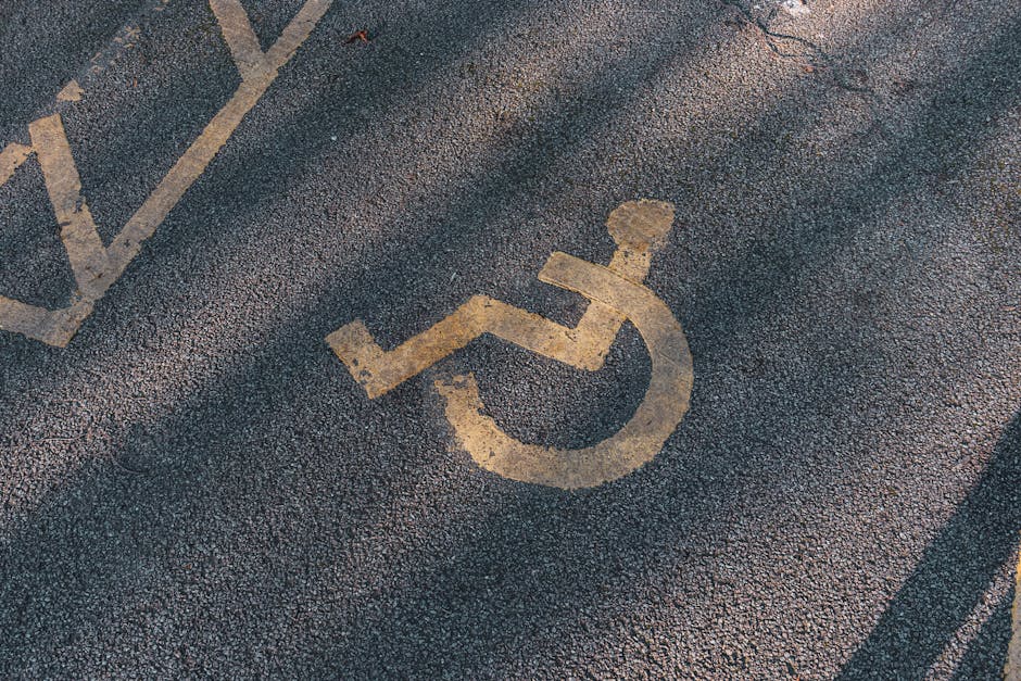 A designated parking space with a yellow painted wheelchair symbol on dark asphalt, situated next to a building entrance with a narrow pathway leading to a doorway. The parking area includes white lin