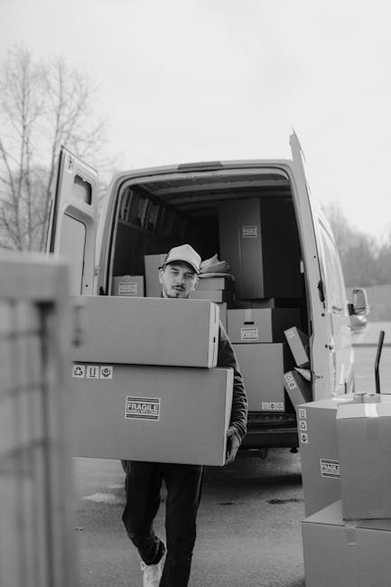 A male removal specialist from Removal Van Merton is seen loading cardboard boxes into the back of a white van parked on a cobblestone street in an urban residential area during daytime. The open carg
