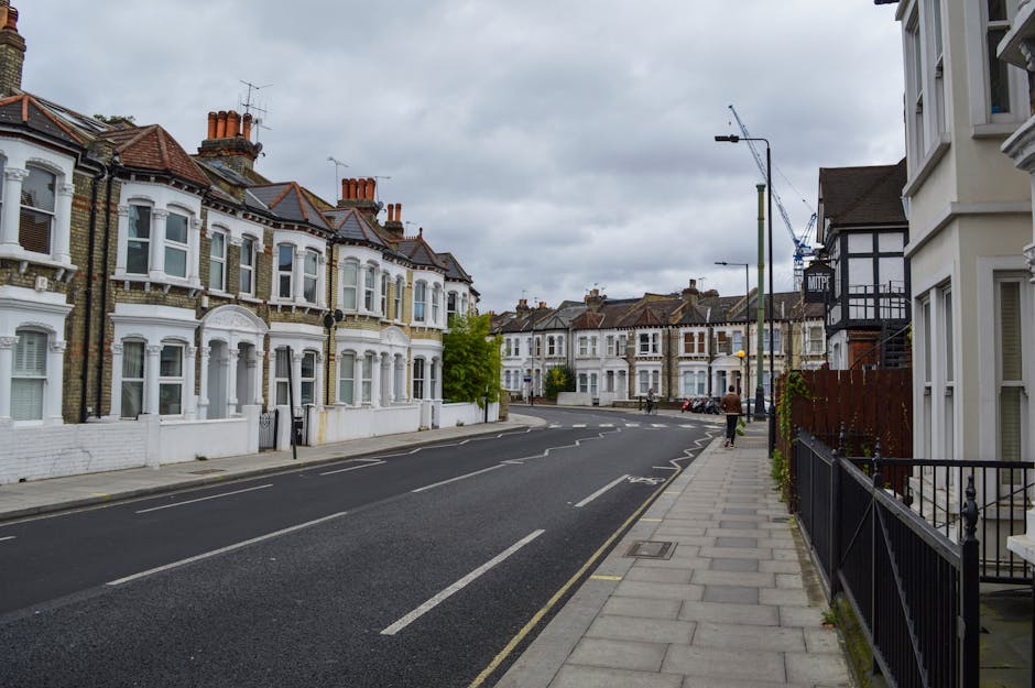 A wide, quiet residential street in Wimbledon Village, Merton, showing a row of Victorian-style terraced houses with bay windows, brickwork facades, and chimneys, under an overcast sky. The street features a paved sidewalk with metal railings, a few parked cars, and a walking pedestrian. In the foreground, a removal van from Removal Van Merton is parked close to one of the houses, ready for a home relocation or furniture transport. Visible on the pavement are cardboard boxes, plastic-wrapped furniture, and blankets, indicating packing activities for a move. The scene captures the loading process of household items into the vehicle, with some objects on the driveway near the building entrance, and a trolley nearby. Construction cranes in the background suggest ongoing developments in the area, while streetlights and trees add to the typical suburban atmosphere fitting for a house removals service in Merton.
