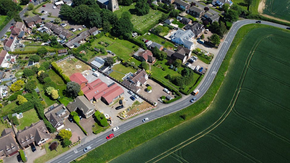 A wide, quiet residential street in Wimbledon Village, Merton, showing a row of Victorian-style terraced houses with bay windows, brickwork facades, and chimneys, under an overcast sky. The street features a paved sidewalk with metal railings, a few parked cars, and a walking pedestrian. In the foreground, a removal van from Removal Van Merton is parked close to one of the houses, ready for a home relocation or furniture transport. Visible on the pavement are cardboard boxes, plastic-wrapped furniture, and blankets, indicating packing activities for a move. The scene captures the loading process of household items into the vehicle, with some objects on the driveway near the building entrance, and a trolley nearby. Construction cranes in the background suggest ongoing developments in the area, while streetlights and trees add to the typical suburban atmosphere fitting for a house removals service in Merton.