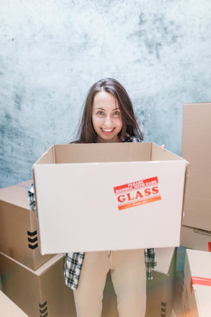 A woman with long brown hair and a smile is standing indoors, holding an open cardboard box labeled 'GLASS' in red text and reinforced with packing tape, as part of a home relocation process associated with house removals services. Behind her, there are additional cardboard boxes stacked on the floor for packing and moving preparations. The background features a plain, light blue wall with a slightly textured finish, indicating an interior environment suitable for packing items before loading into a removal van. The scene reflects the packing stage of furniture transport and moving logistics, with the woman preparing fragile items for transport, supported by Removal Van Merton's professional removals services.
